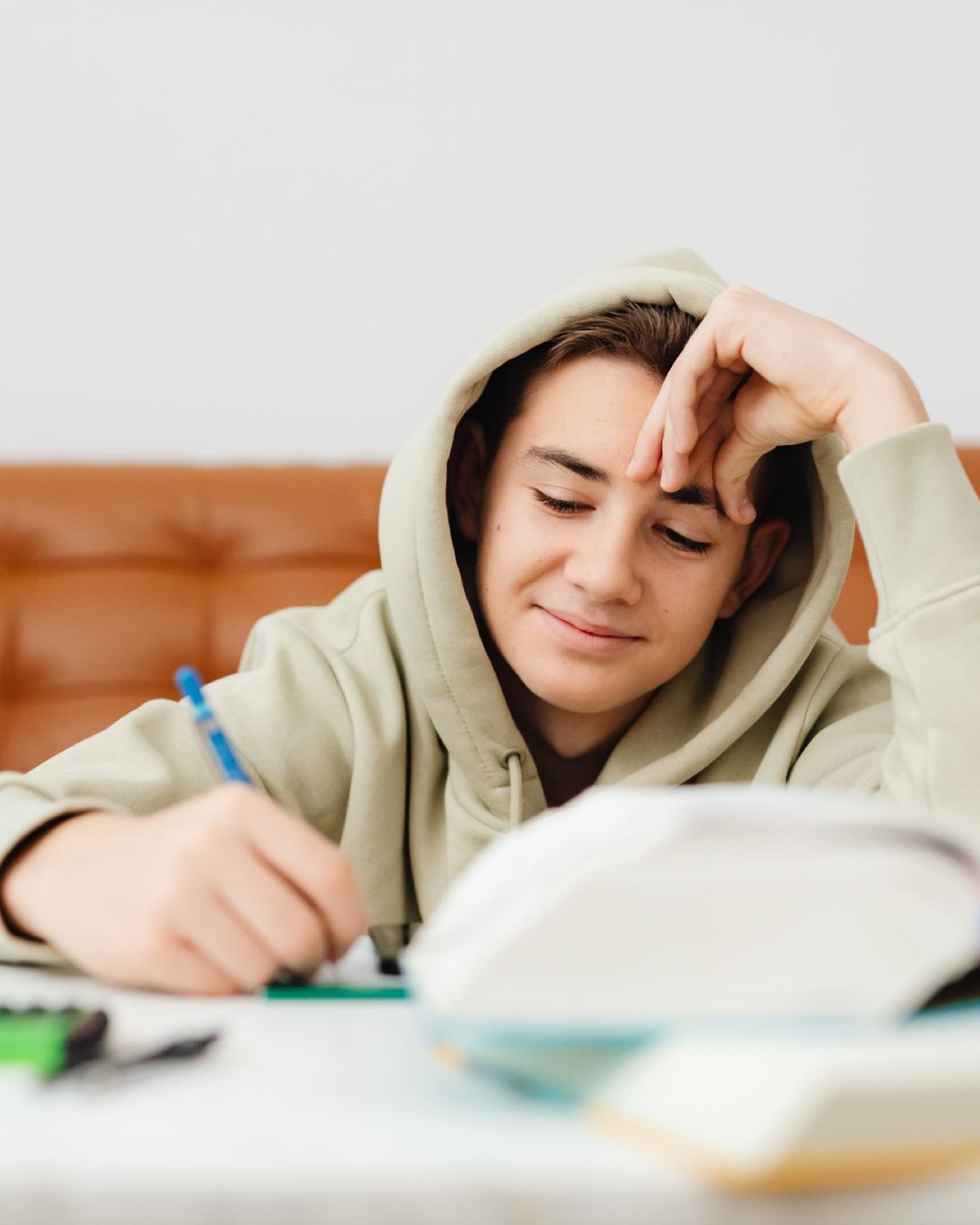 A teenage boy in a hoodie smiling as he writes notes at a table with open books