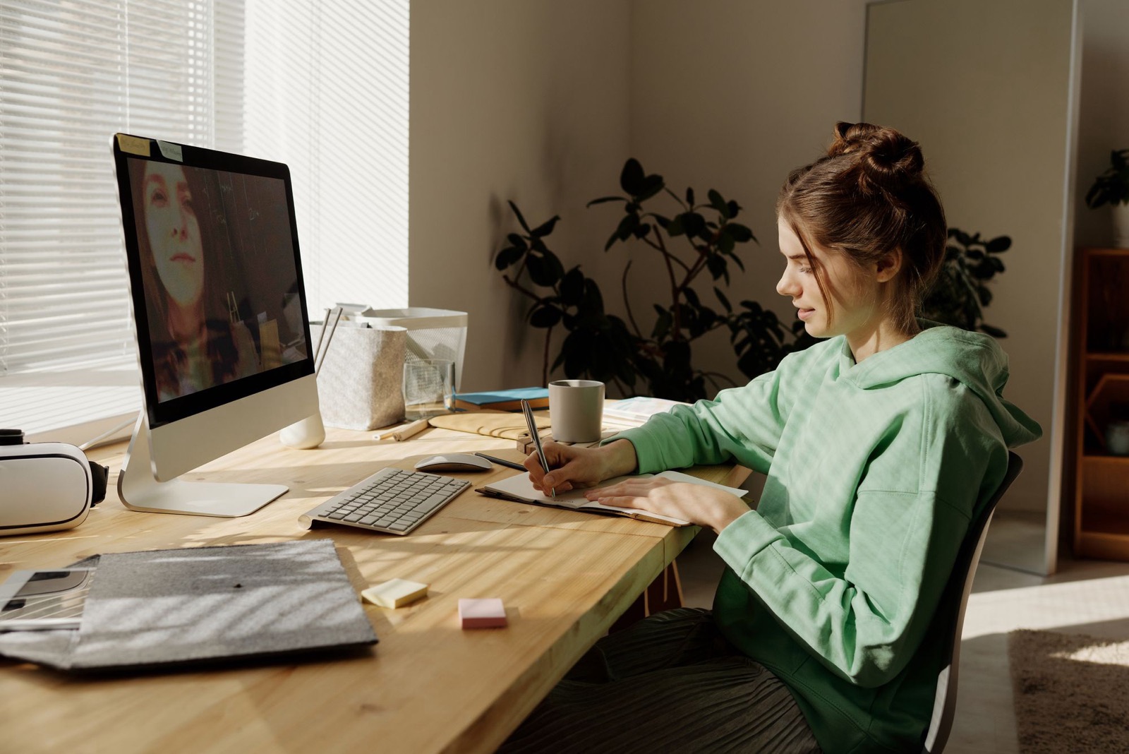 A teenage girl taking notes at a sunny desk during a video call on a desktop computer