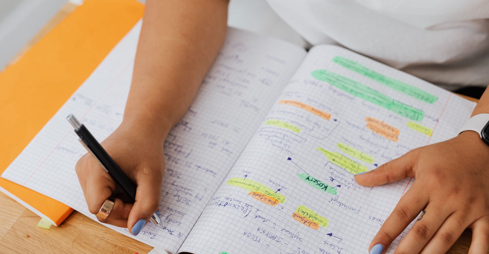 Hands writing colour-highlighted revision notes in a squared notebook on a wooden desk
